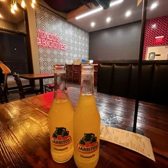Two bottles of pineapple Jarritos on a wooden table under the Tacos El Bigotes neon sign