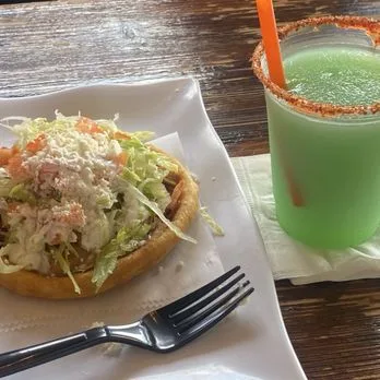 Sope topped with lettuce and cotija next to a green frozen margarita with a chile rim